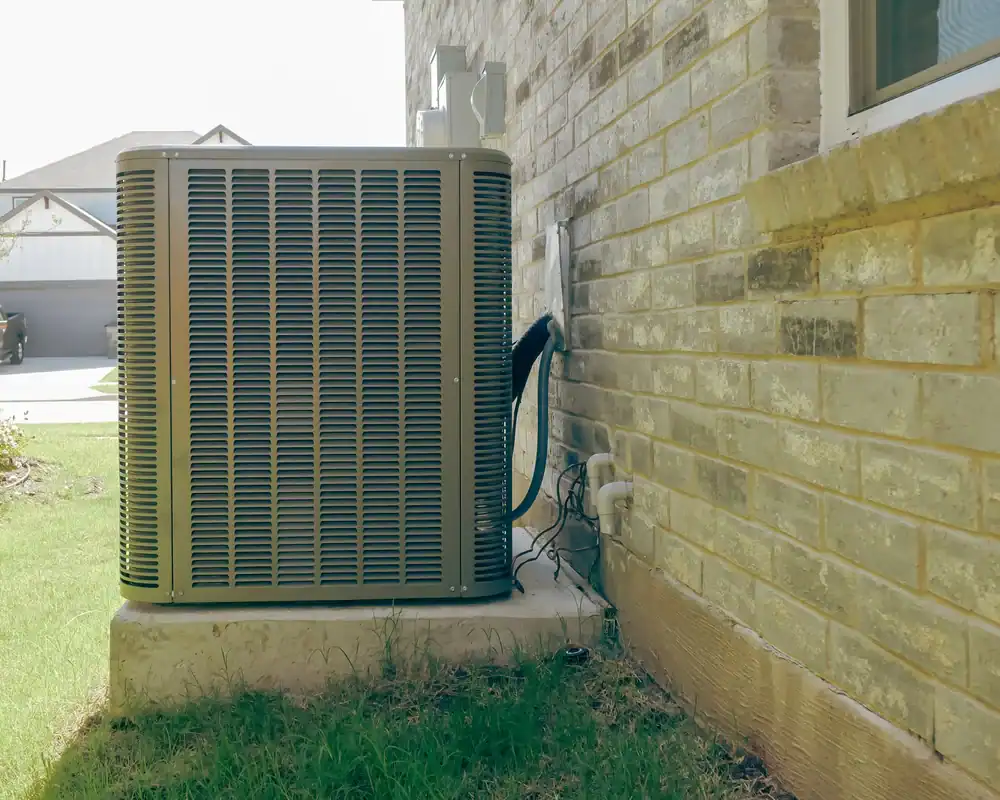 An outdoor air conditioning unit sits on a concrete slab next to a brick house, with grass growing nearby and cables connecting the unit to the building—expertly installed by HVAC contractor services New York City.