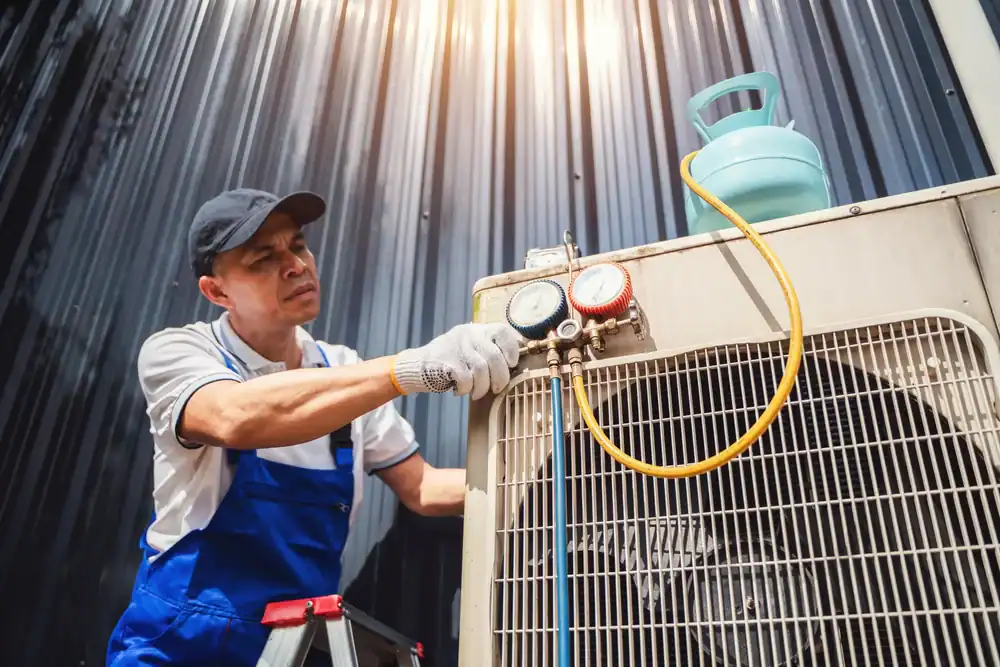 A technician in a blue uniform and cap checks an air conditioner unit with pressure gauges and hoses outside, showcasing reliable HVAC contractor services New York City under the bright sunlight.