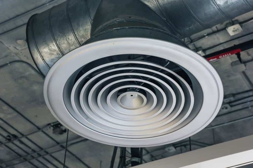 Close-up view of a round white air vent or diffuser attached to an exposed metal air duct on a ceiling, showcasing the quality of HVAC contractor services New York City with visible pipes and wiring in the background.