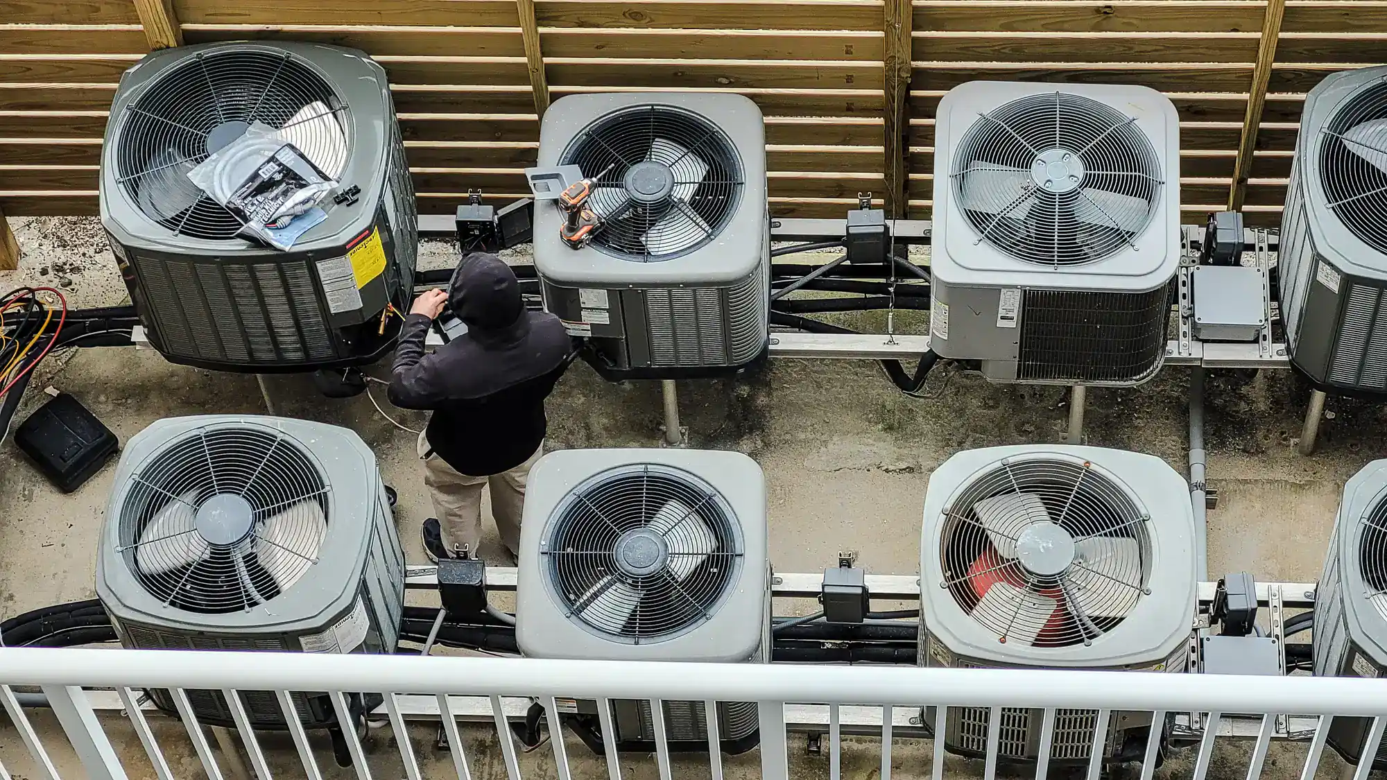 A person in a hoodie is working on a row of six outdoor air conditioning units, using tools and diagnostic equipment. The units are lined up beside a wooden fence, seen from above.