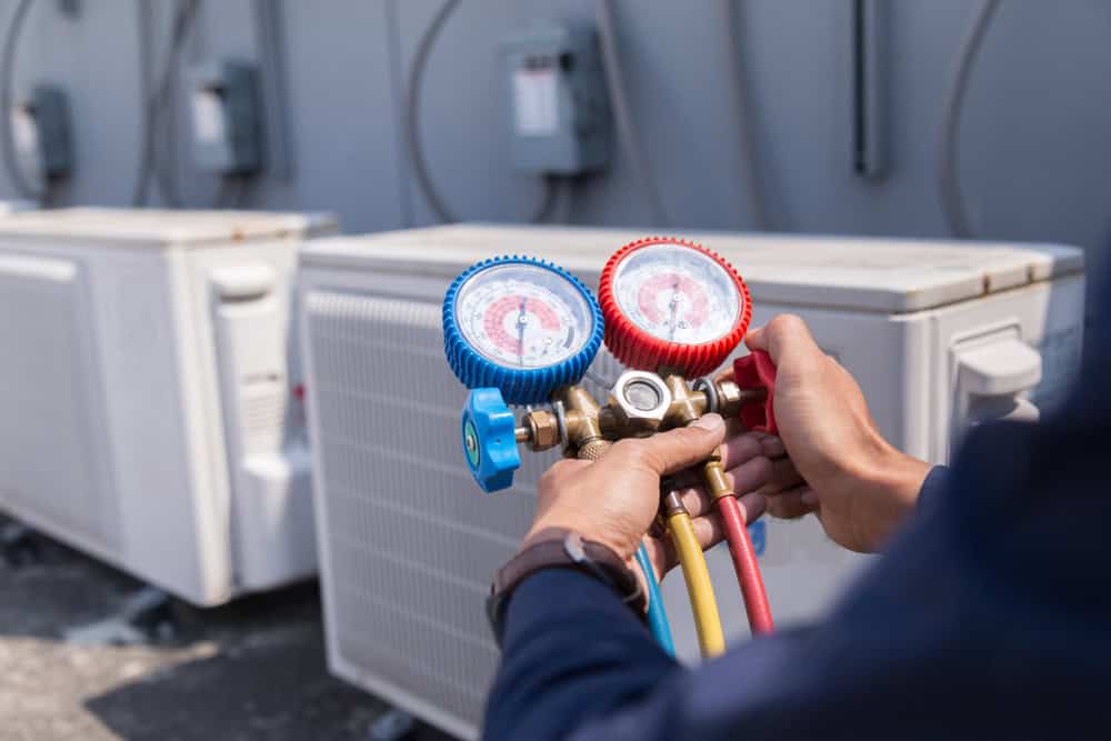 A person holds HVAC manifold gauges to check air conditioning units outdoors, with several AC units visible in the background.